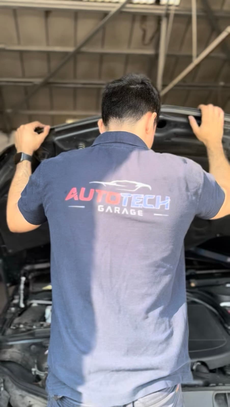 A technician leaning over the open bonnet of a vehicle, seen from behind, working in the engine bay.