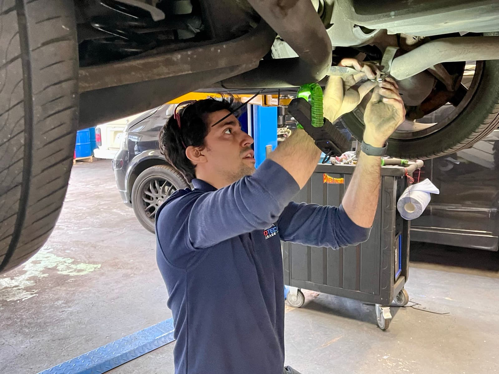 A technician working beneath a vehicle raised on the workshop ramp, holding an inspection lamp.
