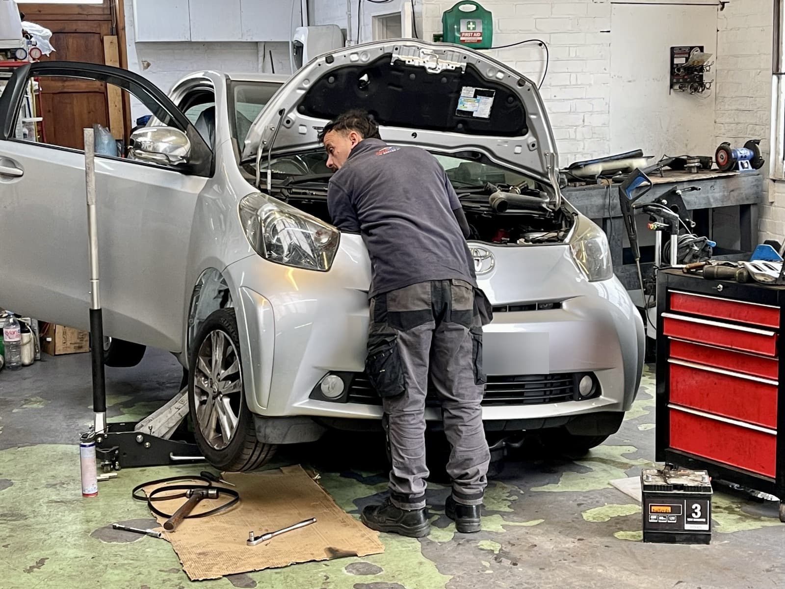 A technician leaning into the engine bay of a small silver hatchback, bonnet raised, inside the workshop.